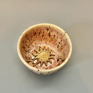 May include: A small ceramic bowl with a speckled brown and pink glaze. The bowl features a raised gold sun design in the centre.