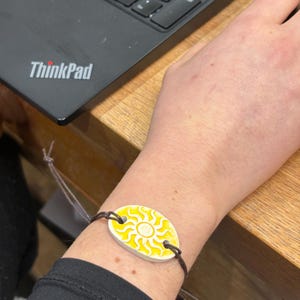 May include: An oval bracelet featuring a yellow sun design on a white background. The bracelet is attached to a brown cord. The image shows the bracelet on a wrist. The background includes a laptop.