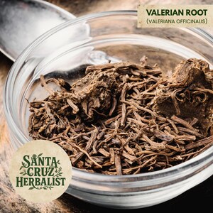 Puede incluir: Un cuenco de vidrio transparente lleno de raíz de valeriana seca, con el texto "Valerian Root (Valeriana officinalis)" encima. El cuenco está sobre una superficie de madera y se ve el logotipo de Santa Cruz Herbalist.