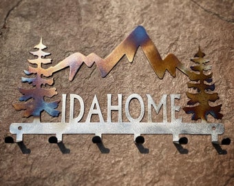 May include: Metal wall art featuring the word "IDAHOME" with a mountain and tree design. The piece has a silver finish with hints of blue and gold. Includes six hooks for hanging items.