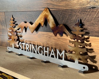 May include: Metal wall art featuring a mountain range and pine trees design. The word "STRINGHAM" is cut out below the mountains. The piece has a silver finish with brown and gold accents. Includes hooks for hanging.