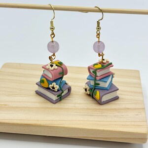 May include: A pair of dangle earrings featuring a stack of three books with floral details and a light purple bead at the top.