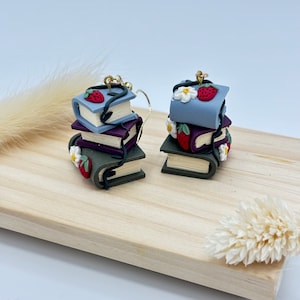 Puede incluir: Un par de pendientes hechos a mano de arcilla polimérica con pilas de libros en miniatura con detalles florales y fresas. Los libros son en tonos de azul, morado, verde y negro.