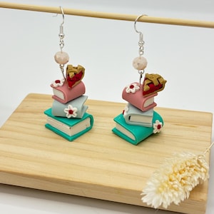 May include: A pair of dangle earrings featuring a stack of four miniature books topped with a cherry pie slice. The books are pink, light blue, and teal. The earrings have silver hooks and white flower accents.