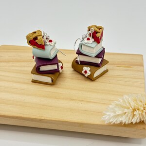 May include: A pair of whimsical earrings featuring stacks of miniature books in brown, purple, and light blue. Each stack is topped with a slice of pie and adorned with white flower accents. The earrings are displayed on a wooden board.