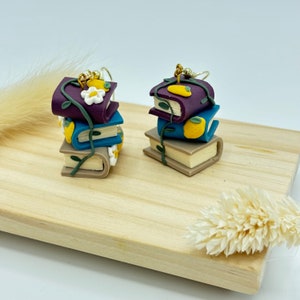 May include: A pair of dangle earrings featuring a stack of four miniature books with vines and flowers. The books are purple, blue, brown, and beige. The earrings are made of polymer clay.