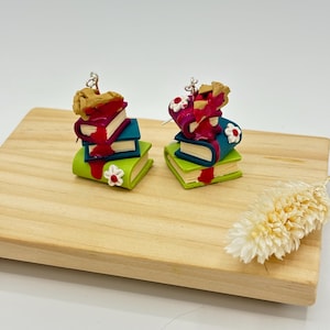 May include: A pair of dangle earrings featuring a stack of books with a pie on top. The books are green, teal, and purple with red accents. The pie is brown with a red filling. The earrings have silver hooks.