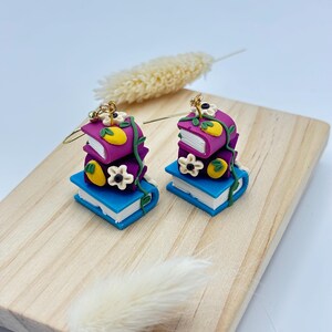 May include: A pair of dangle earrings featuring a stack of three books with floral and lemon accents. The books are purple, blue, and white with gold accents. The earrings are made of polymer clay.