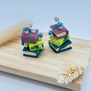 May include: A pair of earrings featuring miniature stacks of books with blue and white mushrooms on top. The books are green, purple, and blue.