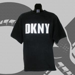 May include: Black DKNY t-shirt with white block lettering. The short-sleeved shirt is displayed on a mannequin. The background is a grayscale design. The shirt is made of a soft material.