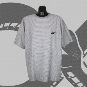 Puede incluir: Una camiseta a rayas grises y blancas con mangas cortas. La camiseta tiene un pequeño logotipo en el pecho. La camiseta se muestra en un maniquí negro. El fondo es un diseño gris y blanco.