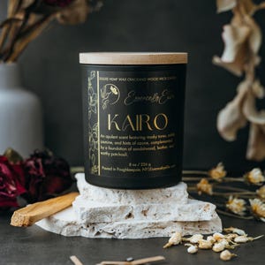 May include: Black candle jar with a wooden lid and gold lettering. The candle is labeled "Essence B Ellie KAIRO" and describes the scent as "an opulent scent featuring musky tones, subtle jasmine, and hints of ozone, complemented by a foundation of sandalwood, leather, and earthy patchouli." The candle is 8 oz / 226 g and poured in Poughkeepsie, NY.