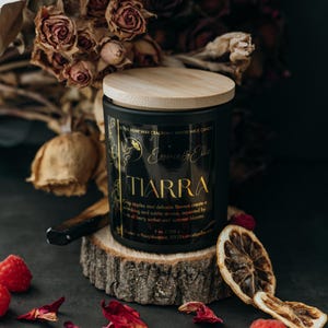 May include: A black candle jar with a wooden lid. The candle is labeled "Essence by Ellie" and "TIARRA". The label also describes the scent as "Crisp apples and delicate flowers create a refreshing and subtle aroma, accented by hints of berry sorbet and summer blooms."