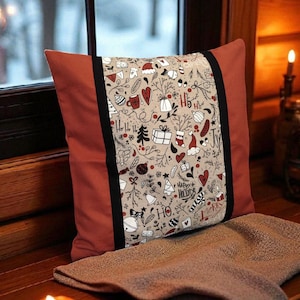Könnte beinhalten: Ein dekoratives Kissen mit festlichem Weihnachtsdesign. Das Kissen hat ein zentrales Feld mit weihnachtlichen Illustrationen in Rot, Schwarz und Weiß auf beigem Hintergrund. Die Seiten sind einfarbig rostfarben, mit einem schwarzen Streifen, der die Felder trennt.