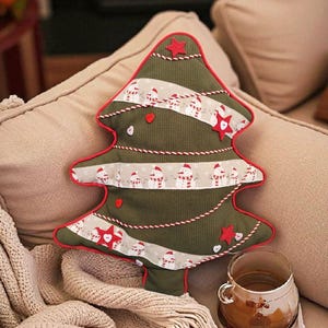 May include: A Christmas tree-shaped pillow with a red border, green body, and festive snowman and star accents. The pillow is on a beige couch with a cup of coffee and a cozy blanket.