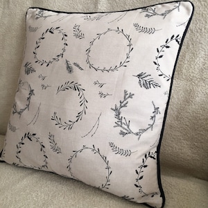 May include: Square decorative pillow with a cream-colored background and a black botanical wreath pattern. The pillow has a black border. The design features various wreath and sprig illustrations.