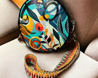 May include: A round, black crossbody bag with a colorful abstract floral print. The bag has a long, colorful woven strap.