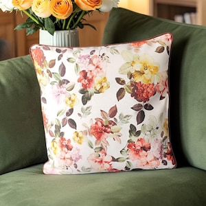 May include: A square decorative pillow with a floral pattern featuring red, yellow, and pink flowers on a white background. The pillow is placed on a green sofa, with a vase of orange and white roses in the background.