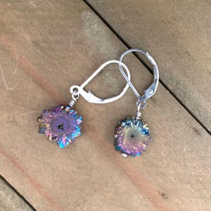 May include: Earrings with iridescent druzy stone pendants. The pendants have a mix of purple, blue, and gold colours, with a central cavity. The earrings are suspended from silver-coloured leverback closures. The background is a wooden surface.