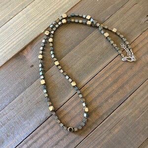 May include: A necklace featuring a repeating pattern of small, metallic dark grey and polished gold-tone cube-shaped beads. The strand includes a silver-tone metal lobster claw clasp and an adjustable chain link extender at the end of the necklace.