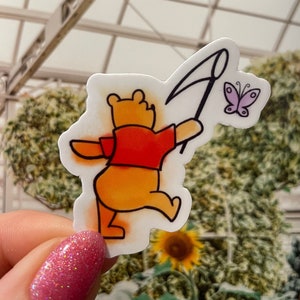 May include: A Winnie the Pooh sticker with a red shirt and a net in his hand, chasing a purple butterfly. The sticker is on a white background.