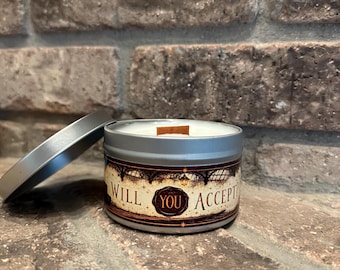 May include: A round silver metal candle tin with a wooden wick and a label featuring a dark, ornate architectural design. The label displays the text "WILL YOU ACCEPT?" in a stylized font, centered around a circular wax seal graphic.