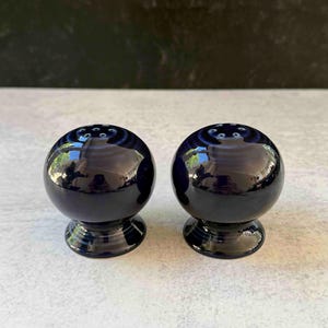 May include: Two matching, spherical, navy blue salt and pepper shakers. Each shaker has a glossy finish and sits on a small, pedestal base. The top of each shaker has several small holes for dispensing.