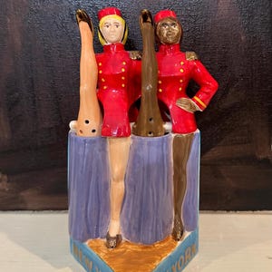 Puede incluir: Figura de cerámica que representa a dos botones con uniformes rojos y detalles dorados. Los botones están sobre una base azul con la inscripción "New York" en dorado. La figura tiene una estética vintage.