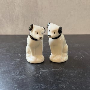 Puede incluir: Se muestran dos figuras de perros de cerámica, probablemente saleros y pimenteros. Los perros son blancos con detalles negros en las orejas y collares. Están sentados, con un acabado brillante, sobre una superficie oscura.