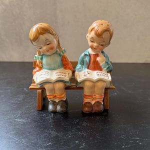 May include: A pair of ceramic salt and pepper shakers depicting children reading from books while seated on a wooden bench. The figures are dressed in colorful clothing, with detailed facial features and hair.