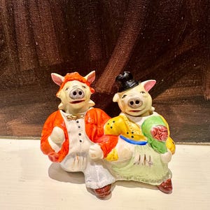 Puede incluir: Un par de figuras de cerdos de cerámica, una con una chaqueta naranja y la otra con un vestido amarillo. Las figuras tienen orejas rosas y llevan sombreros. El fondo es marrón oscuro.