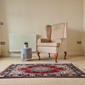 Puede incluir: Un sillón de orejas beige con patas de madera se encuentra sobre una alfombra beige con una alfombra estampada en rojo, negro y crema delante de él. Un pequeño taburete gris con patas doradas se encuentra al lado del sillón.