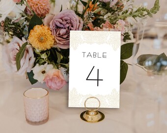 May include: A table number card with the word "TABLE" and the number "4" in black font. The card is white with a decorative cream-colored border. A gold-colored metal holder supports the card. A bouquet of flowers is in the background.