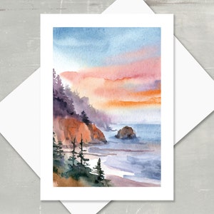 May include: Watercolor painting of a coastal landscape with a sunset. The artwork features a rocky coastline, ocean, and a sky with shades of blue, orange, and pink. The painting is on a white card, with a white envelope.
