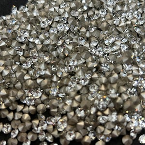 Op de afbeelding: Een close-up van een grote hoeveelheid zilveren en heldere strass steentjes. De strass steentjes zijn verspreid over een donker oppervlak.
