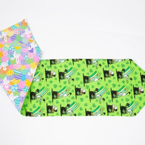 May include: A green table runner with a repeating pattern of a cartoon leprechaun wearing a hat and holding a pot of gold. The runner is trimmed with a pink and white fabric with a pattern of Easter eggs and bunnies.