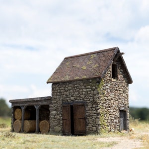 Puede incluir: Un pequeño granero de piedra con techo de paja y una puerta de madera. Hay tres balas de heno delante del granero.