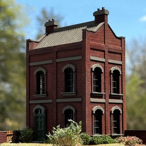 May include: A red brick building with multiple windows and a dark green door. The building has a grey roof and is surrounded by green bushes.