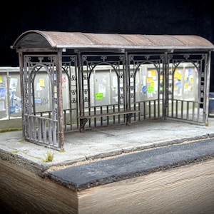 Peut inclure: Maquette de miniature d'un arrêt de bus avec une structure métallique vieillie, un banc et un toit. La scène comprend un tableau d'affichage avec des affiches colorées et une poubelle. Le tout est posé sur une base en béton avec une bande d'asphalte.
