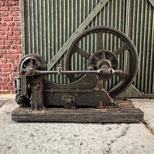 Op de afbeelding: Een model van een stationaire motor van donker metaal met een groot vliegwiel met spaken en een kleiner riemschijfje verbonden door een horizontale stang. Het mechanische geheel is gemonteerd op een rechthoekige houten basis met textuur.