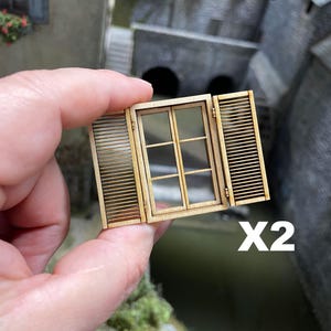 Può includere: Una finestra in miniatura di legno con due persiane, tenuta in mano. La finestra ha un vetro trasparente e un motivo a griglia. Le persiane sono aperte, rivelando un design a lamelle. Il testo "X2" è visibile.