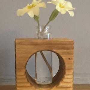 May include: A wooden vase with a circular cutout to hold a small glass bottle with white flowers. The vase is made of natural wood with a rustic finish.