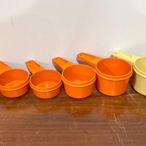 May include: Set of five orange and yellow plastic measuring cups. The cups are arranged in order of size, with the largest cup on the right and the smallest cup on the left.