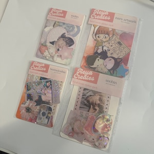 May include: Four packs of hand-made sticker packs with different designs. Each pack features a different artist's work, including illustrations of animals, people, and objects. The packs are labeled with the artist's name and the phrase "Reya Creates".