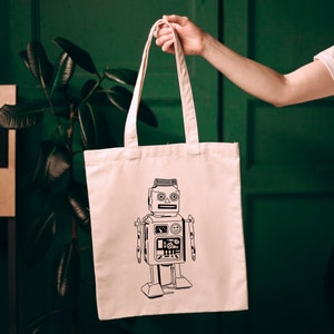 May include: A white tote bag with a black and white illustration of a robot on the front. The robot has a round head with two large eyes and a small mouth. It has a rectangular body with a few details on it. The bag has two handles.