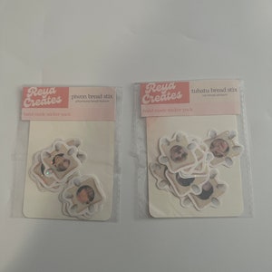 May include: Two clear plastic bags containing sticker packs. Each pack has a white card with pink text that reads "Reya Creates" and the name of the sticker pack. The sticker packs are titled "piwon bread stix" and "tubatu bread stix". The stickers are white with images of bread.
