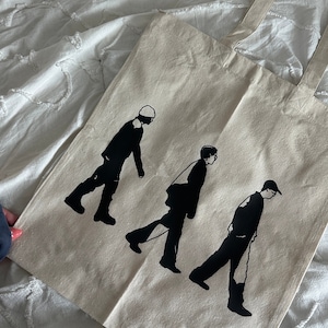 May include: A white canvas tote bag featuring black silhouettes of three people walking. The figures are wearing hats and walking in a line.