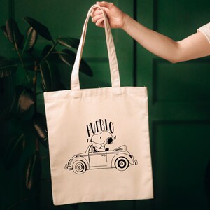 May include: A white canvas tote bag with black handles. The bag features a black and white illustration of Snoopy driving a Volkswagen Beetle with the word "PUEBLO" printed above it.