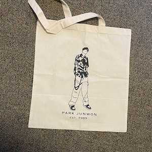 May include: A white canvas tote bag with black handles and a black graphic of a person wearing a patterned shirt and pants. The text "PARK JUNWON EST. 1989" is printed below the graphic.