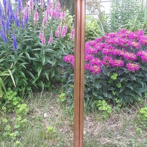 May include: A copper bird bath with a wide, shallow bowl and a tall, slender stand. The stand has three legs that extend out from the base. The bird bath is surrounded by green grass and pink and purple flowers.